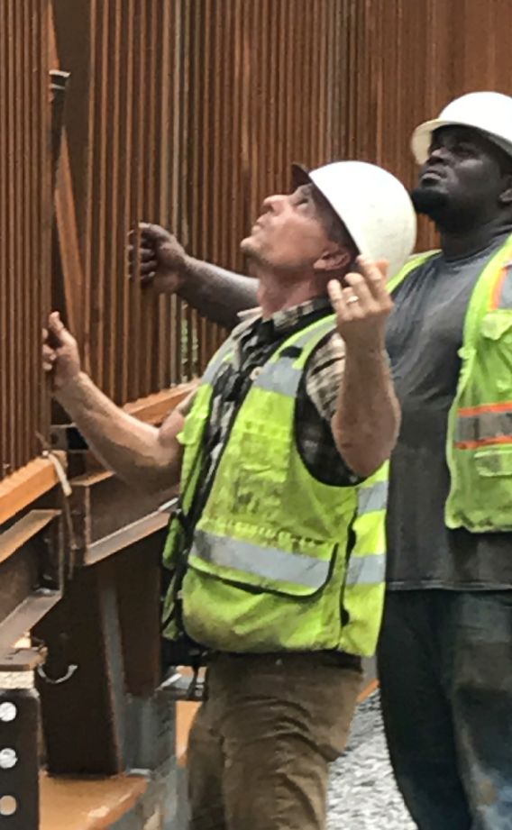 A highway construction worker stands next to a fellow construction worker and both are looking up at a project
